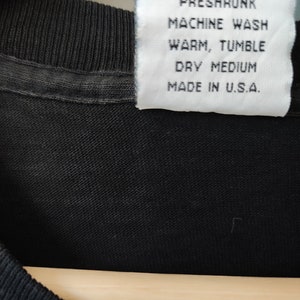 May include: Close-up of a black t-shirt with a white label. The label reads "100% Cotton, Preshrunk, Machine Wash, Warm, Tumble, Dry Medium, Made in U.S.A."