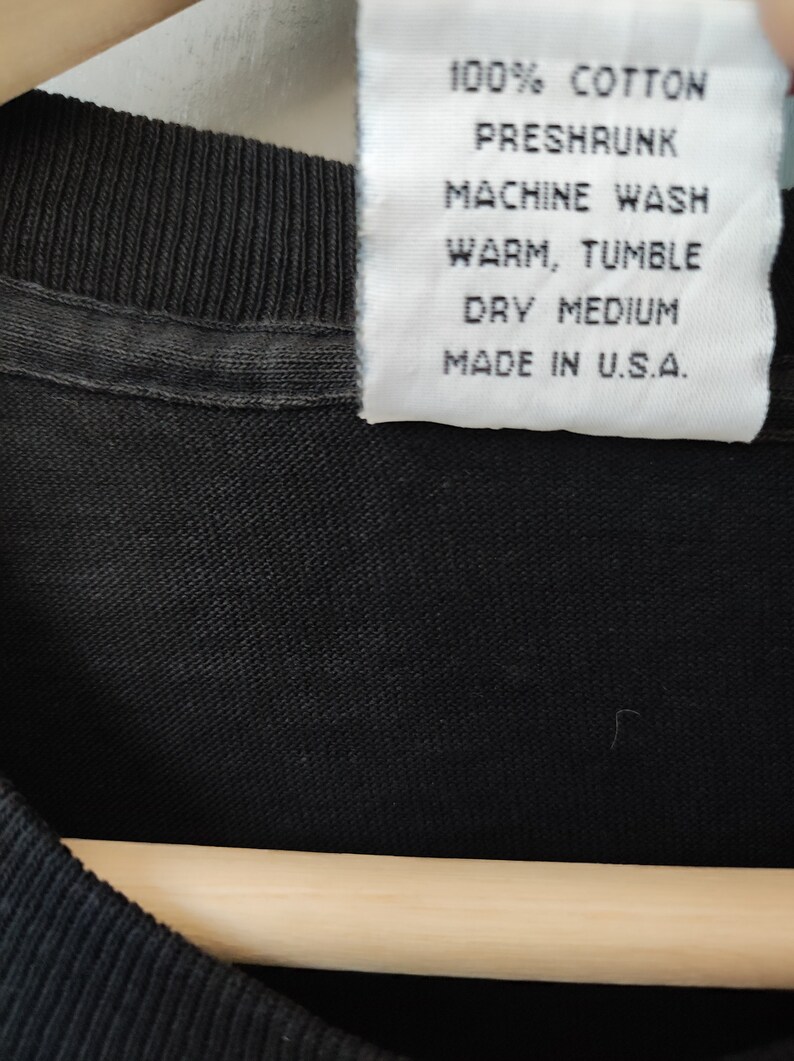 May include: Close-up of a black t-shirt with a white label. The label reads "100% Cotton, Preshrunk, Machine Wash, Warm, Tumble, Dry Medium, Made in U.S.A."
