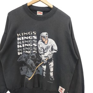 K&ouml;nnte beinhalten: Dunkelgraues Vintage-Sweatshirt mit einer Hockey-Spieler-Grafik und dem Wort "KINGS" vertikal wiederholt. Die Grafik zeigt einen Hockeyspieler in einem wei&szlig;-violetten Trikot. Das Sweatshirt h&auml;ngt an einem Holzb&uuml;gel.