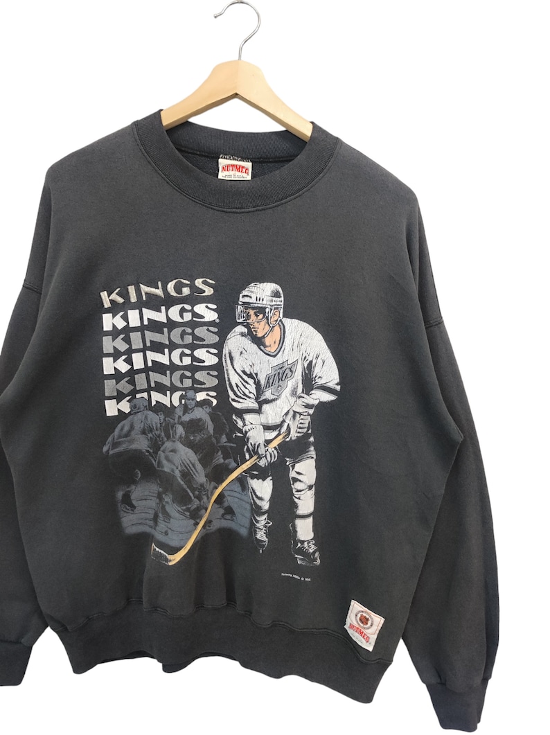 K&ouml;nnte beinhalten: Dunkelgraues Vintage-Sweatshirt mit einer Hockey-Spieler-Grafik und dem Wort "KINGS" vertikal wiederholt. Die Grafik zeigt einen Hockeyspieler in einem wei&szlig;-violetten Trikot. Das Sweatshirt h&auml;ngt an einem Holzb&uuml;gel.