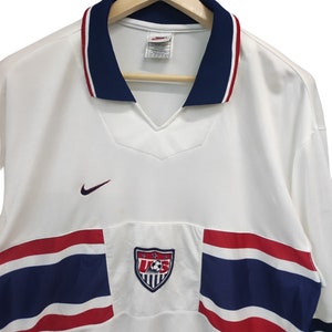 May include: White football shirt with a navy blue collar and red and white trim. Features a U.S. Soccer crest and a Nike logo. The shirt has a V-neck design and short sleeves.