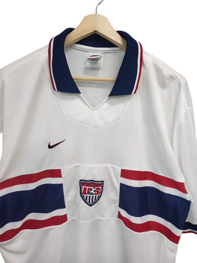 May include: White football shirt with a navy blue collar and red and white trim. Features a U.S. Soccer crest and a Nike logo. The shirt has a V-neck design and short sleeves.