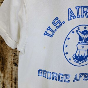 May include: White short-sleeved sweatshirt with blue text and graphics. The text includes "U.S. AIR FORCE" and "GEORGE AFB". A circular graphic with stars and an eagle is also visible. The sweatshirt has a crew neck and short sleeves.