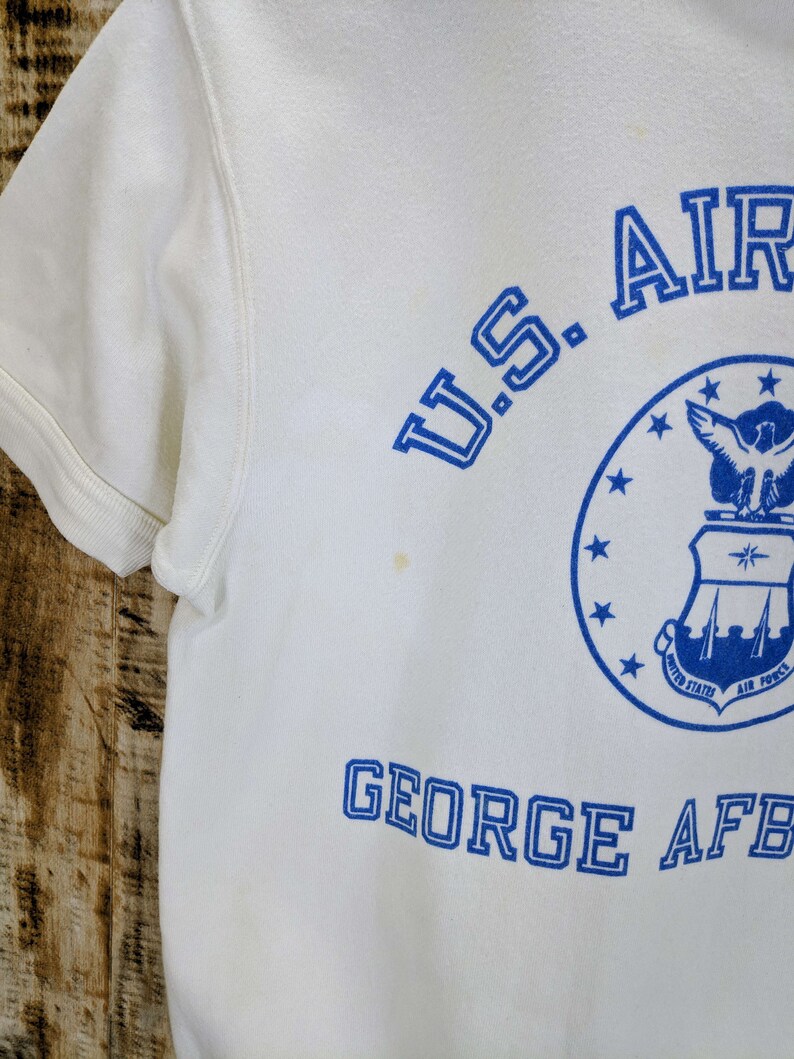 May include: White short-sleeved sweatshirt with blue text and graphics. The text includes "U.S. AIR FORCE" and "GEORGE AFB". A circular graphic with stars and an eagle is also visible. The sweatshirt has a crew neck and short sleeves.