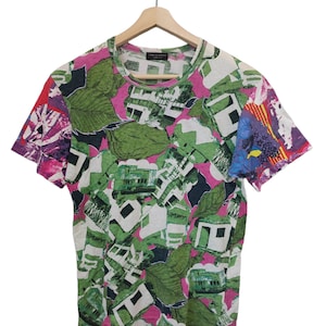 Może przedstawiać: T-shirt z kr&oacute;tkim rękawem z żywym, całopowierzchniowym nadrukiem. Wz&oacute;r przedstawia zielone liście, elementy architektoniczne i abstrakcyjne kształty w kolorach białym, zielonym, r&oacute;żowym i fioletowym. Koszulka wisi na drewnianym wieszaku.