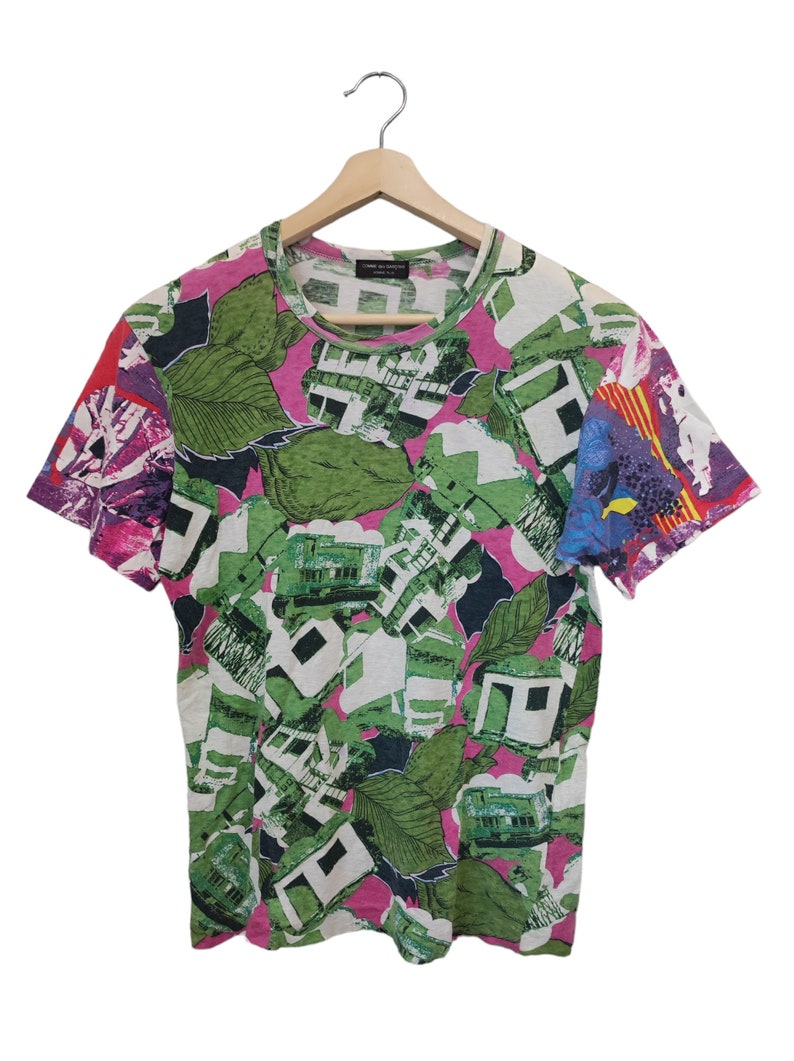 Może przedstawiać: T-shirt z kr&oacute;tkim rękawem z żywym, całopowierzchniowym nadrukiem. Wz&oacute;r przedstawia zielone liście, elementy architektoniczne i abstrakcyjne kształty w kolorach białym, zielonym, r&oacute;żowym i fioletowym. Koszulka wisi na drewnianym wieszaku.