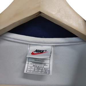May include: A white Nike shirt tag with the Nike logo in red. The tag states "100% Polyester" and "Made in U.S.A." The shirt is on a wooden hanger. The shirt has a navy blue collar. The tag also has the size in French and Spanish.