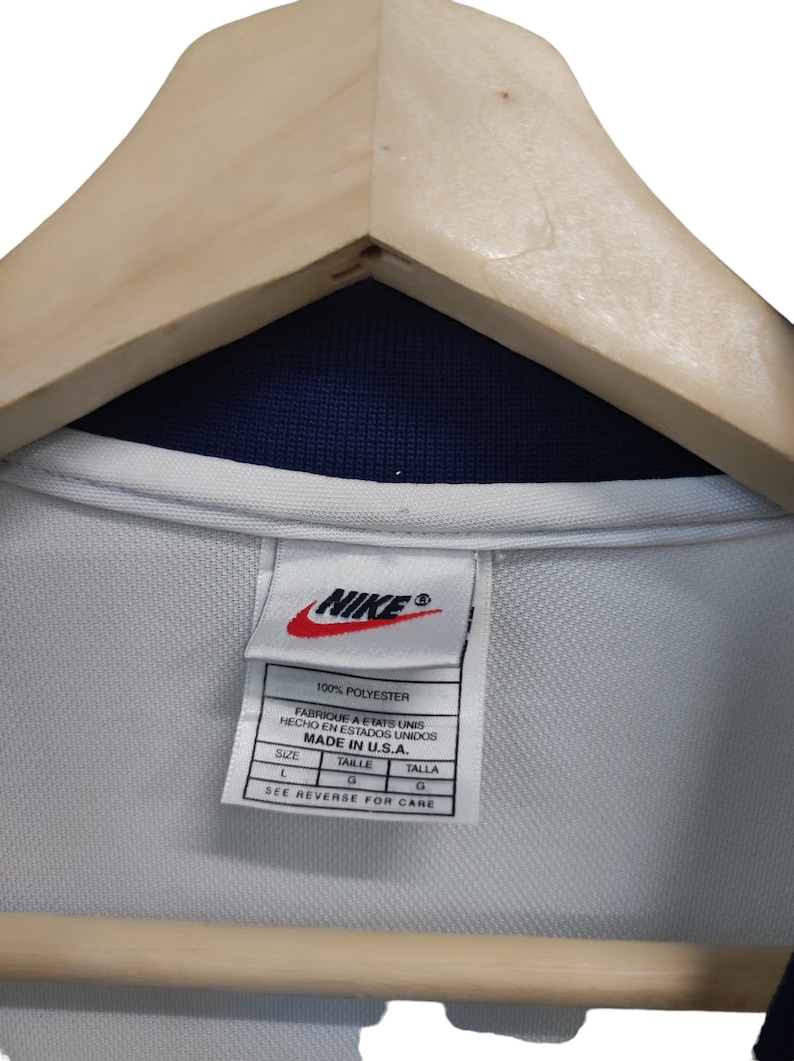 May include: A white Nike shirt tag with the Nike logo in red. The tag states "100% Polyester" and "Made in U.S.A." The shirt is on a wooden hanger. The shirt has a navy blue collar. The tag also has the size in French and Spanish.