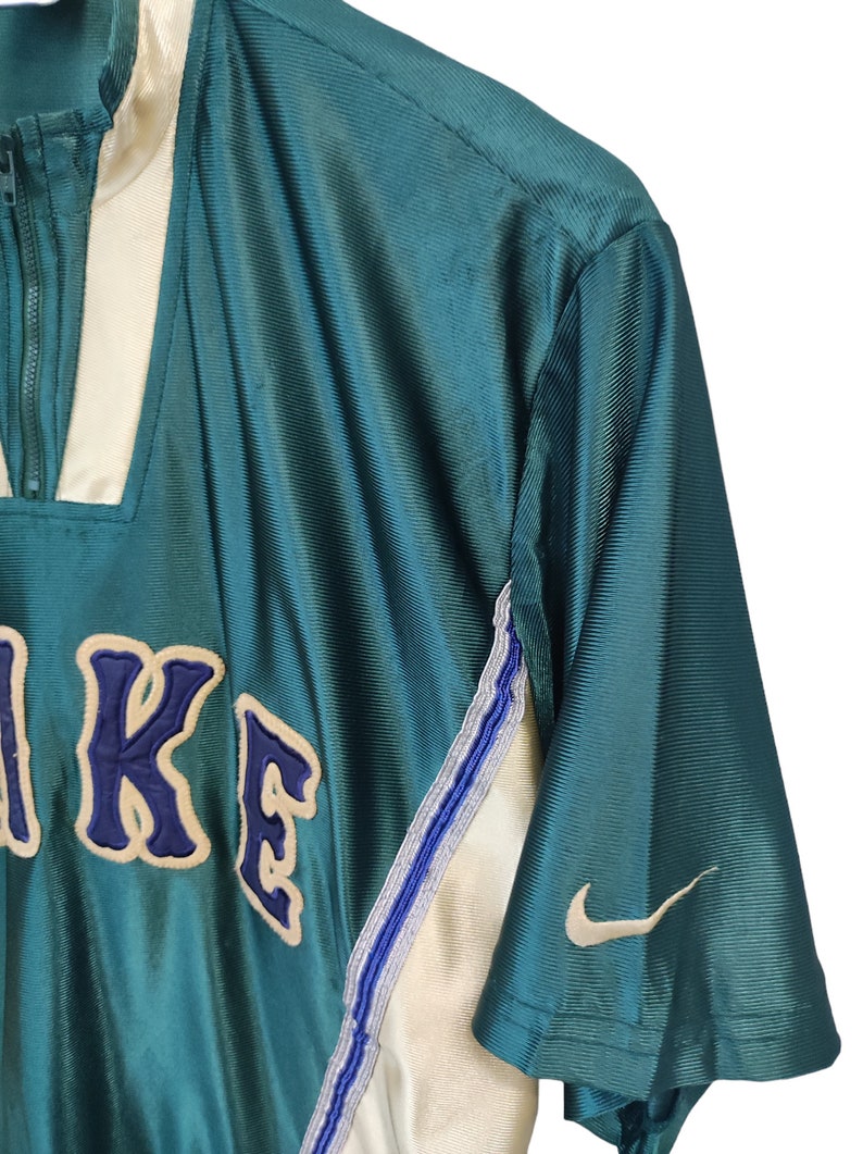 May include: Teal and cream-colored athletic top with a partial zipper and the Nike logo. The top features the Nike name in a blue and cream-colored embroidered design. The sleeve has a white Nike swoosh.