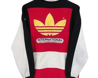 May include: Vintage Adidas International sweatshirt. The sweatshirt features a large gold logo on a red panel, with black and white sleeves and trim. The text "INTERNATIONAL" and "ADIDAS THE WORLD BRAND WITH THREE STRIPES" are printed below the logo.