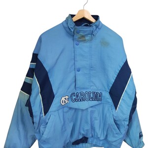 May include: A light blue and navy blue pullover windbreaker jacket featuring the text CAROLINA and a logo on the front. The jacket has a high collar with snap buttons, long sleeves with dark blue panels, and a front pocket with a flap closure.