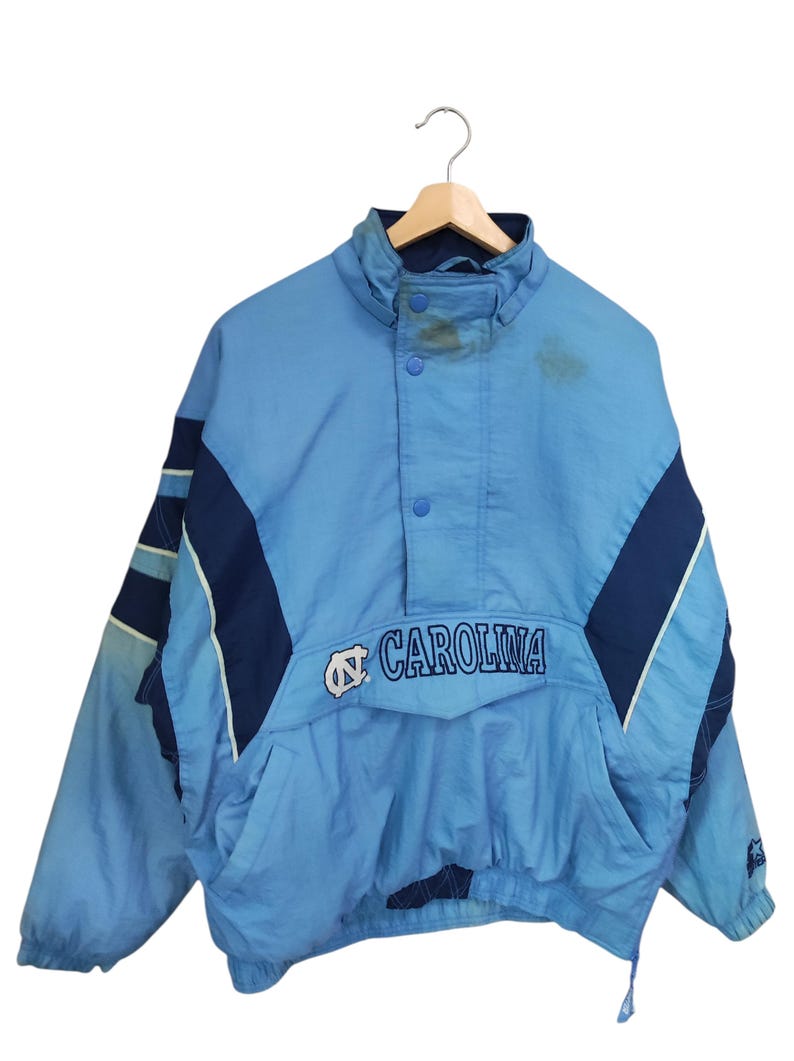 May include: A light blue and navy blue pullover windbreaker jacket featuring the text CAROLINA and a logo on the front. The jacket has a high collar with snap buttons, long sleeves with dark blue panels, and a front pocket with a flap closure.