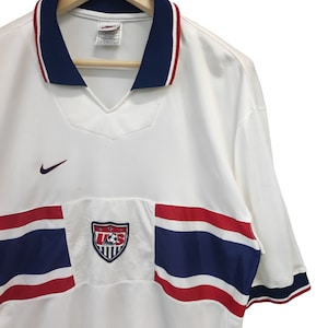 May include: White football shirt with a navy blue collar and red trim. Features the US Soccer crest and a Nike logo. The shirt has red and navy blue stripes across the chest.