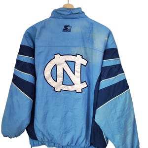 May include: A light blue and navy blue Starter brand windbreaker jacket featuring a large white and navy blue North Carolina Tar Heels logo on the back. The jacket has a high collar, long sleeves with navy accents, and an elasticated hem.