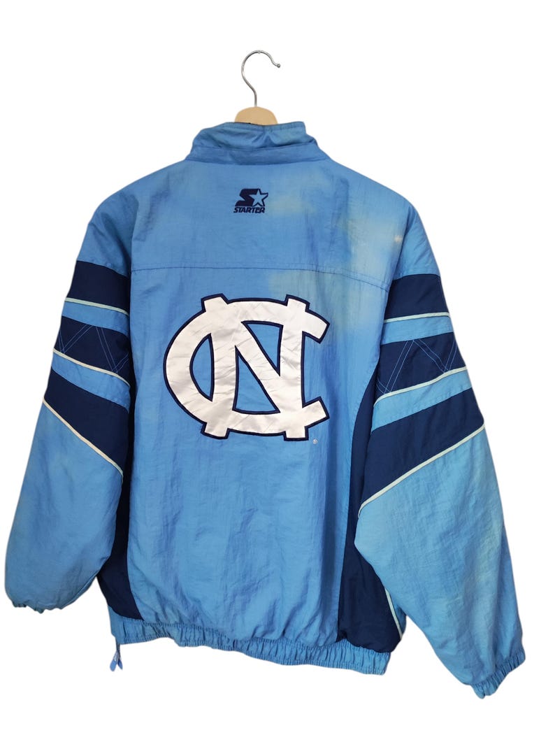 May include: A light blue and navy blue Starter brand windbreaker jacket featuring a large white and navy blue North Carolina Tar Heels logo on the back. The jacket has a high collar, long sleeves with navy accents, and an elasticated hem.