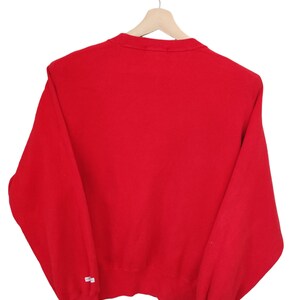 May include: A solid red sweatshirt hangs on a wooden hanger against a white background. The long-sleeved top has a crew neck and ribbed cuffs. A small white tag is visible on the left sleeve. The garment is made of a soft, comfortable material.