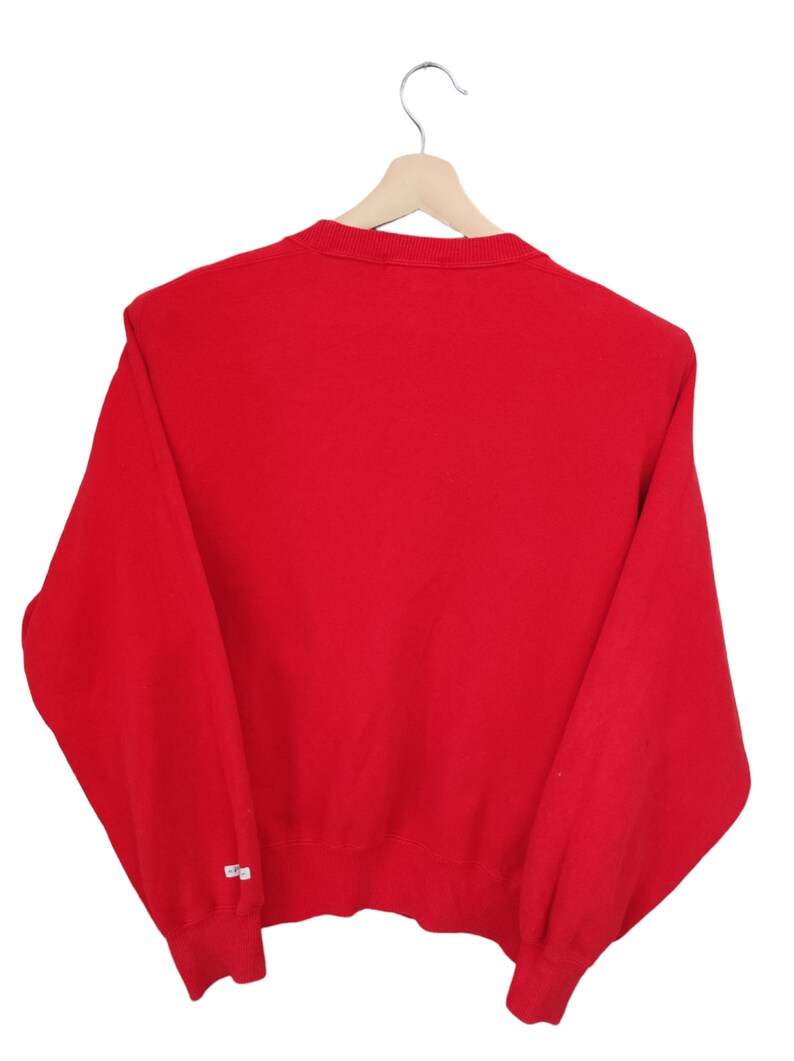May include: A solid red sweatshirt hangs on a wooden hanger against a white background. The long-sleeved top has a crew neck and ribbed cuffs. A small white tag is visible on the left sleeve. The garment is made of a soft, comfortable material.
