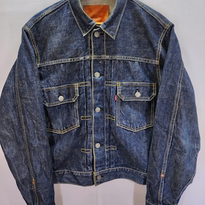 Op de afbeelding: Een donkerblauwe spijkerjas in vintage-stijl, met een klassieke kraag, knoopsluiting en twee borstzakken. De jas heeft een versleten uiterlijk met zichtbare stiksels en metalen knopen. Een klein rood label is zichtbaar op de borstzak.