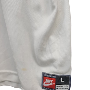May include: Close-up of a white Nike sports jersey. The jersey has a textured, breathable fabric. A Nike label with red and white text is visible, along with a size "L" tag. The jersey has a small stain.