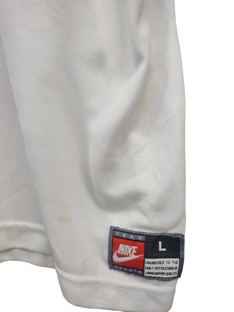 May include: Close-up of a white Nike sports jersey. The jersey has a textured, breathable fabric. A Nike label with red and white text is visible, along with a size "L" tag. The jersey has a small stain.