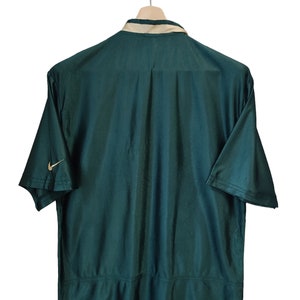 May include: Dark green short-sleeved shirt with a beige collar and the Nike logo on the sleeve. The shirt is hanging on a wooden hanger against a white background. The back of the shirt is visible.