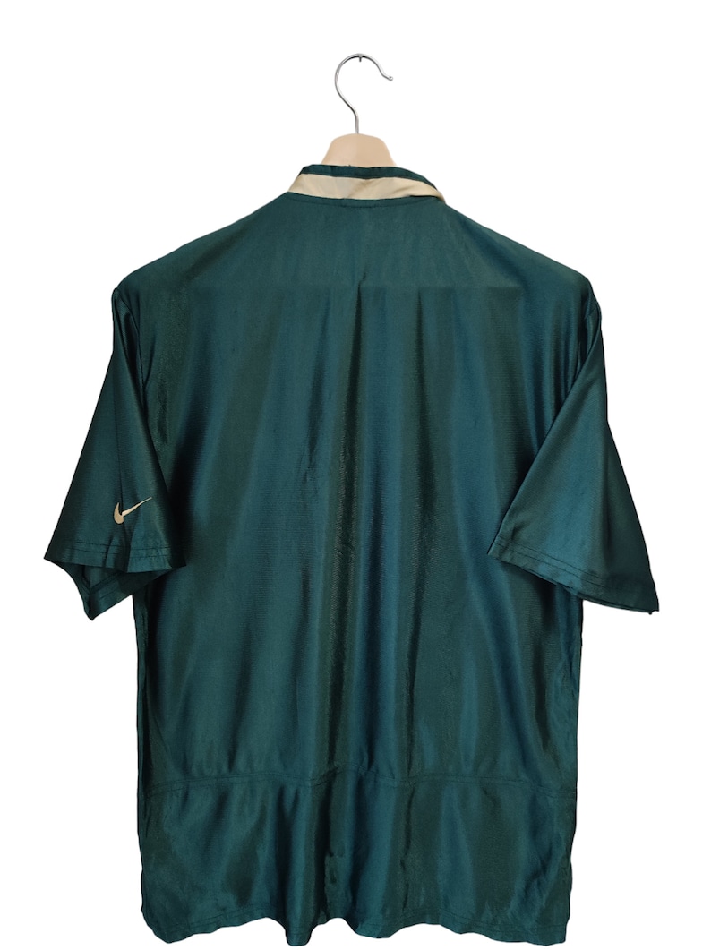 May include: Dark green short-sleeved shirt with a beige collar and the Nike logo on the sleeve. The shirt is hanging on a wooden hanger against a white background. The back of the shirt is visible.