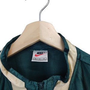 May include: A green and beige Nike track jacket with a full zipper, hanging on a wooden hanger. The Nike logo is visible on a tag. The jacket has beige stripes down the sides and around the collar.