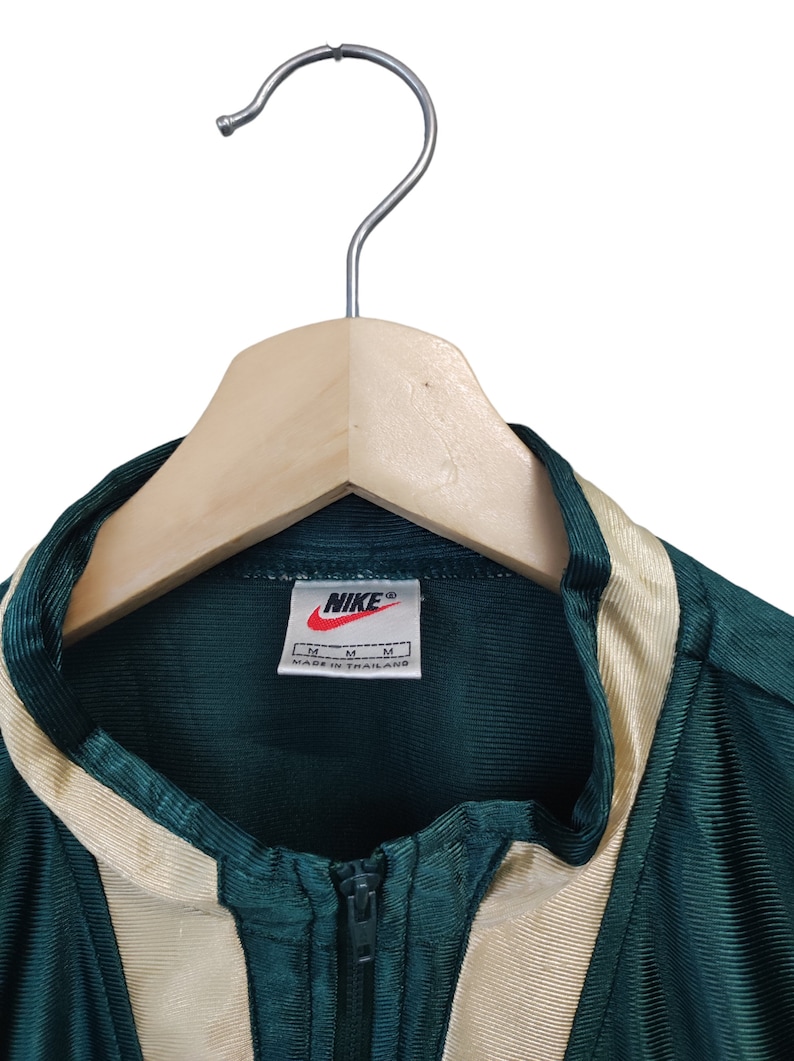 May include: A green and beige Nike track jacket with a full zipper, hanging on a wooden hanger. The Nike logo is visible on a tag. The jacket has beige stripes down the sides and around the collar.