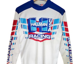 Op de afbeelding: Wit race-shirt met blauwe en rode details. De tekst "HALLMAN RACING USA" staat in een vetgedrukte lettertype op de borst. De mouwen hebben een herhalend patroon van blauwe en rode vierkanten. De kraag en manchetten zijn blauw.