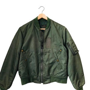 May include: Olive green bomber jacket with a ribbed collar, cuffs, and hem. Features a zip-up front, multiple pockets, and a small patch on the chest. The jacket is hanging on a wooden hanger.