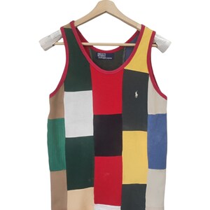 May include: A patchwork tank top with a variety of colors, including red, green, yellow, and blue. The tank top has a red trim and a small logo of a polo player. The tank top is hanging on a wooden hanger.