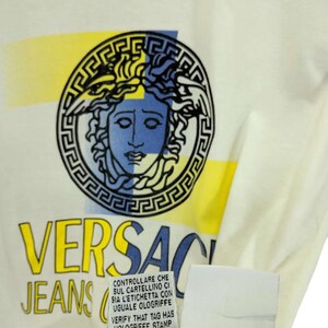 May include: Cream-colored t-shirt featuring the Versace Jeans logo. The logo includes a Medusa head in a circular frame with yellow and blue accents. The shirt also has a tag with Italian text.