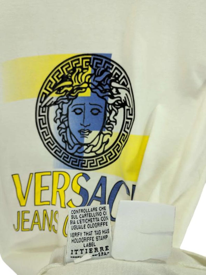 May include: Cream-colored t-shirt featuring the Versace Jeans logo. The logo includes a Medusa head in a circular frame with yellow and blue accents. The shirt also has a tag with Italian text.