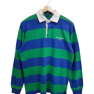 May include: A long-sleeved rugby shirt with horizontal stripes in green and blue. The shirt has a white collar and a three-button placket. The text "POLO SPORT" is visible on the chest, along with a small flag emblem. The shirt is hanging on a wooden hanger.