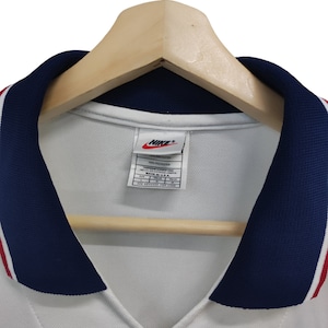 May include: White athletic jersey with a navy blue collar and red and white stripes. The jersey has a V-neck design and a Nike logo on the tag. The jersey is hanging on a wooden hanger.