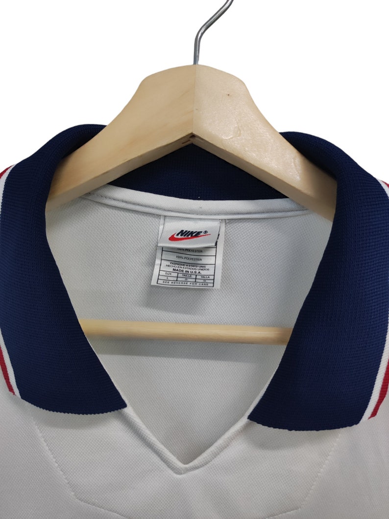 May include: White athletic jersey with a navy blue collar and red and white stripes. The jersey has a V-neck design and a Nike logo on the tag. The jersey is hanging on a wooden hanger.