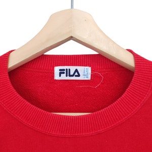 May include: A red crew neck sweatshirt with the Fila logo on a white tag. The sweatshirt is hanging on a wooden hanger. The collar and cuffs are ribbed.