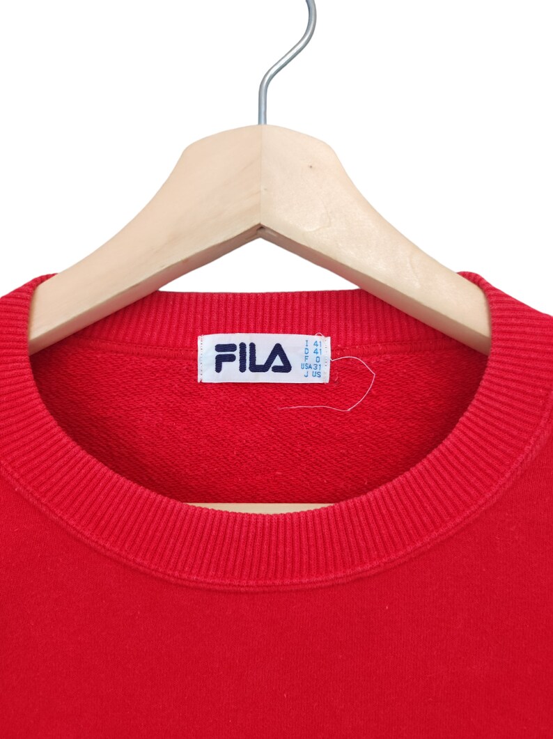 May include: A red crew neck sweatshirt with the Fila logo on a white tag. The sweatshirt is hanging on a wooden hanger. The collar and cuffs are ribbed.