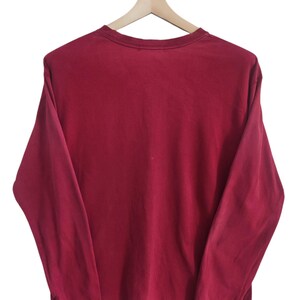 Puede incluir: Una camiseta de manga larga de color granate liso colgada de una percha de madera. La camiseta tiene cuello redondo y un corte holgado. La tela parece ser un material suave y ligeramente descolorido. El fondo es blanco liso.