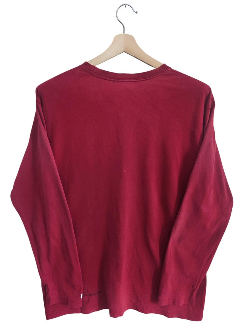 Puede incluir: Una camiseta de manga larga de color granate liso colgada de una percha de madera. La camiseta tiene cuello redondo y un corte holgado. La tela parece ser un material suave y ligeramente descolorido. El fondo es blanco liso.