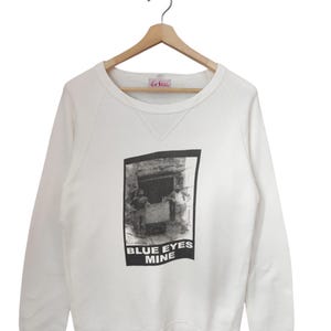 May include: White crew neck sweatshirt with a black and white graphic print. The print features a vintage-style image of people and the text "BLUE EYES MINE". The sweatshirt is hanging on a wooden hanger.