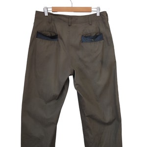 May include: Olive green pants with black pocket accents, hanging on a wooden hanger. The pants have a relaxed fit and a classic design, suitable for casual wear. The back pockets feature a contrasting black material.