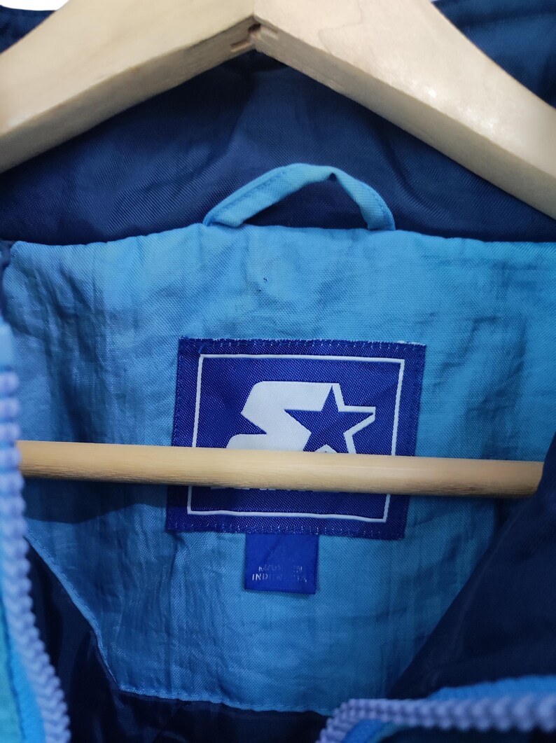 May include: A blue jacket featuring a light blue interior lining and a rectangular Starter brand label in white and blue. The label displays a stylised S logo with a star. A smaller blue tag below the main label indicates the country of origin.