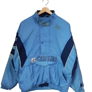 May include: A light blue and navy blue pullover windbreaker jacket featuring the text CAROLINA and a logo on the front chest. The jacket has a high collar with snap buttons, long sleeves with elastic cuffs, and a large front pocket with a flap.