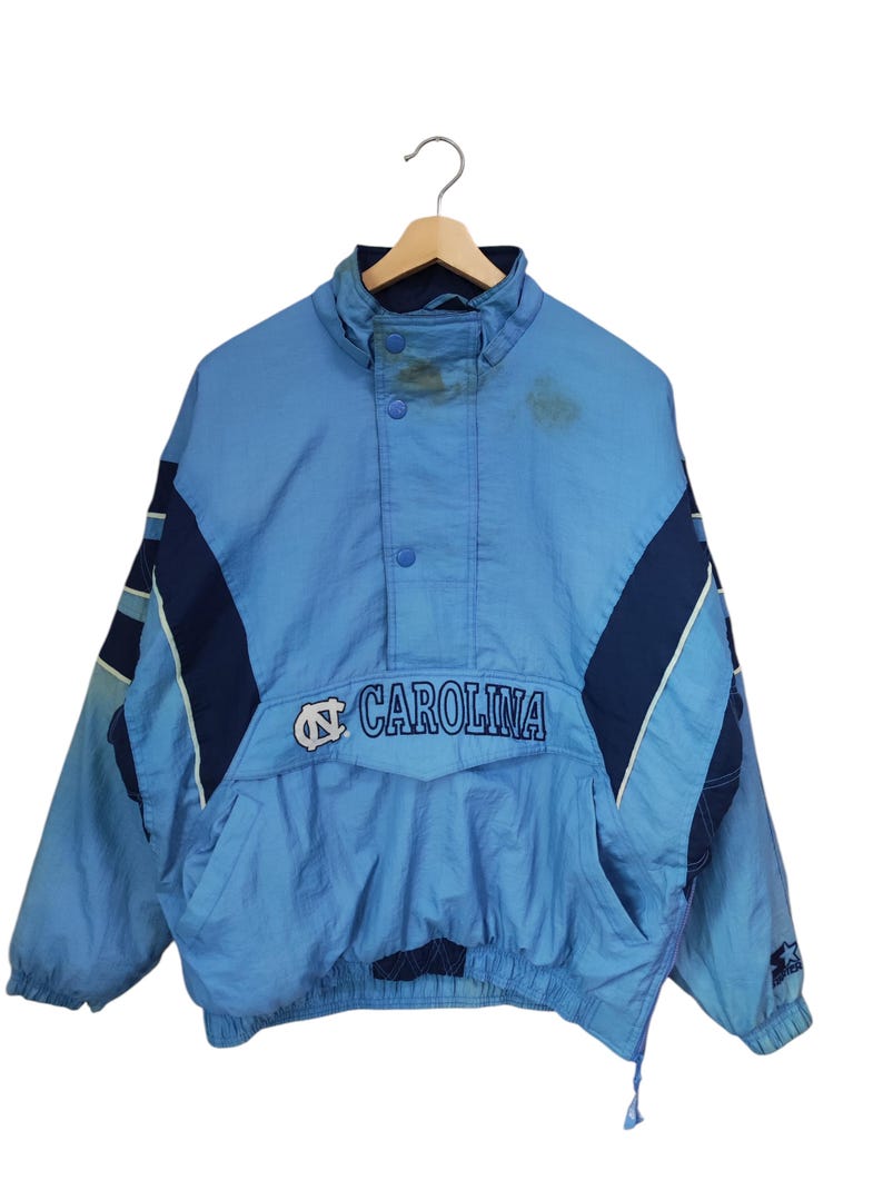 May include: A light blue and navy blue pullover windbreaker jacket featuring the text CAROLINA and a logo on the front chest. The jacket has a high collar with snap buttons, long sleeves with elastic cuffs, and a large front pocket with a flap.