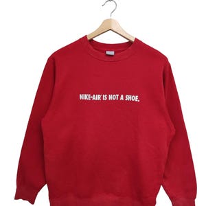 May include: Red crewneck sweatshirt with the text "NIKE-AIR IS NOT A SHOE" in white. The sweatshirt is hanging on a wooden hanger against a white background. A small green Nike logo is visible on the lower left.
