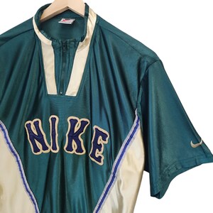 May include: Teal and cream-colored vintage Nike athletic top with a zip-up collar. The word "NIKE" is embroidered across the chest in navy blue with a cream outline. A small Nike swoosh logo is visible on the sleeve.