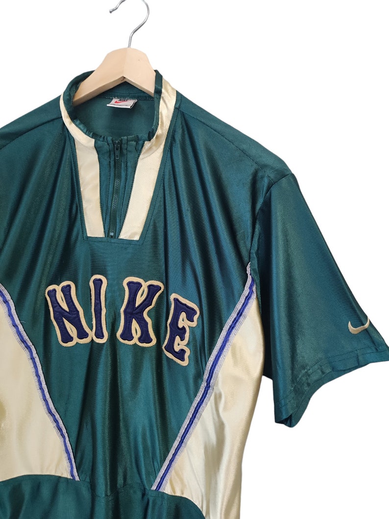 May include: Teal and cream-colored vintage Nike athletic top with a zip-up collar. The word "NIKE" is embroidered across the chest in navy blue with a cream outline. A small Nike swoosh logo is visible on the sleeve.