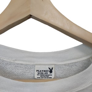 May include: White crew neck sweatshirt with a tag that reads "PLAYBOY M (38-40)". The tag also indicates the material composition: 50% KODEL POLYESTER, 50% COTTON. The sweatshirt is hanging on a wooden hanger.