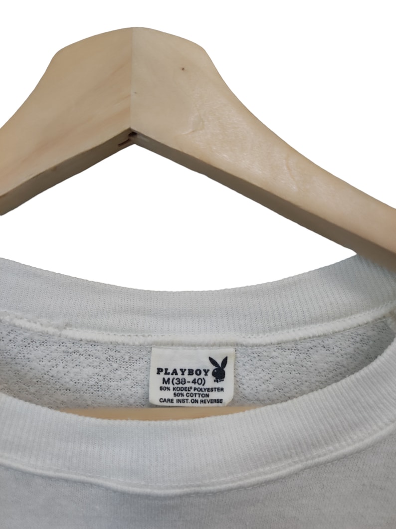 May include: White crew neck sweatshirt with a tag that reads "PLAYBOY M (38-40)". The tag also indicates the material composition: 50% KODEL POLYESTER, 50% COTTON. The sweatshirt is hanging on a wooden hanger.